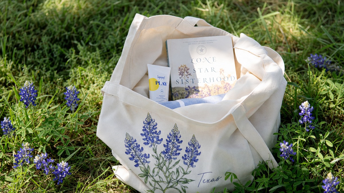 A bluebonnet-print canvas tote bag laid on sun-warmed grass surrounded by fresh bluebonnet flowers in a Texas spring park